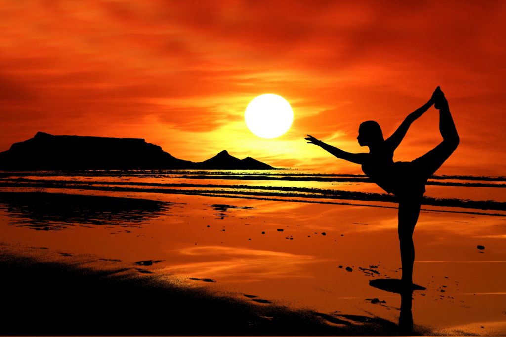 A young girl doing yoga on a beach with sunset and orange yellow clouds